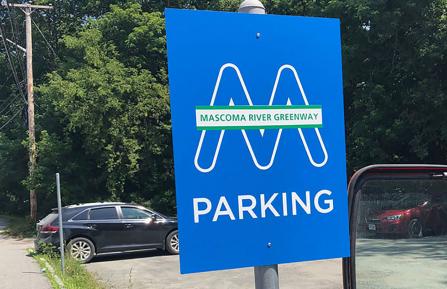 A blue sign that reads Mascoma River Greenway Parking, with a parking lot in the background.